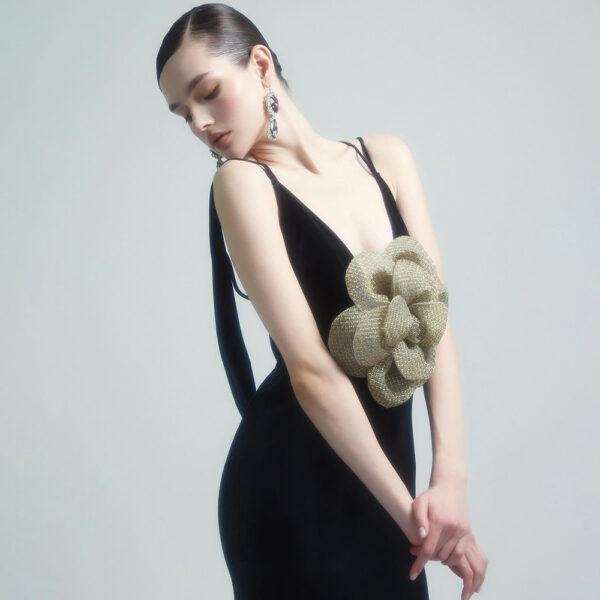 Elegant model showcasing a black evening gown with a large gold flower embellishment, styled with statement earrings and a sleek hairstyle, against a soft gray background.