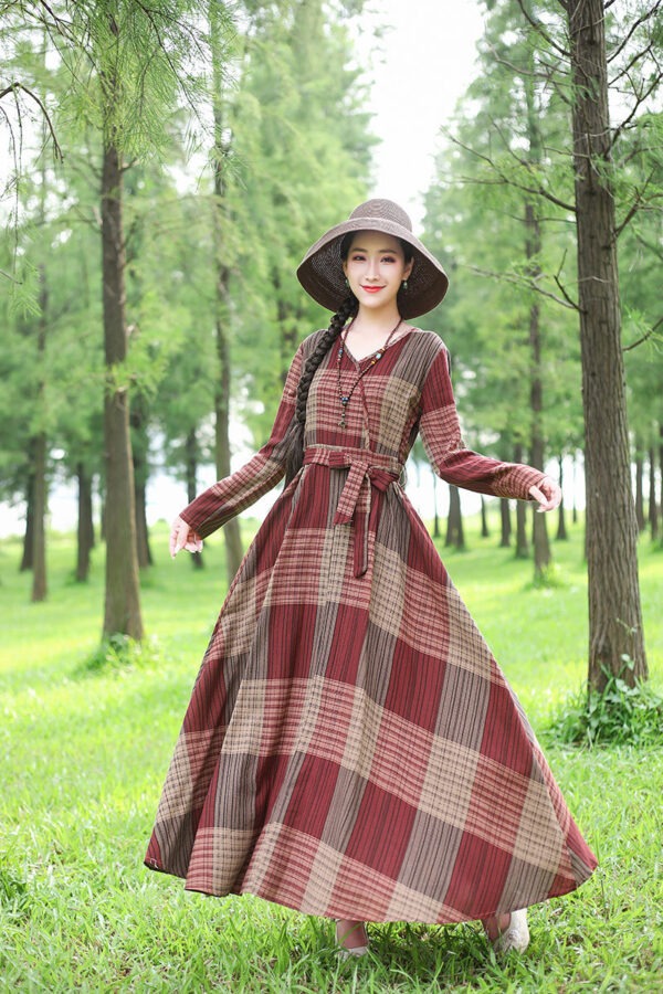 A woman in a long, plaid dress with red, brown, and beige colors twirls in a lush green forest. She wears a wide-brimmed hat and has long braided hair, standing among tall trees in a serene outdoor setting.