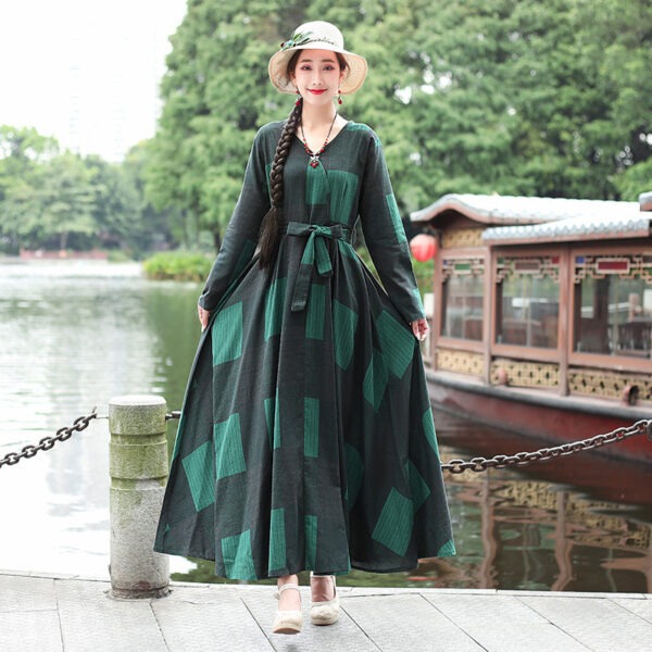 A woman wearing a long, green patterned dress stands by a serene lake, showcasing her stylish outfit and braided hairstyle, with a traditional boat and lush greenery in the background.