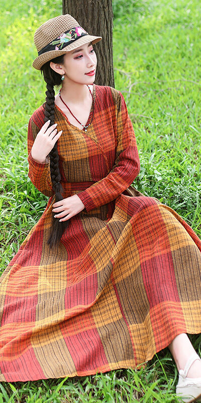 Woman sitting on grass wearing a long plaid dress in warm colors, paired with a straw hat and stylish shoes, enjoying a sunny outdoor setting.