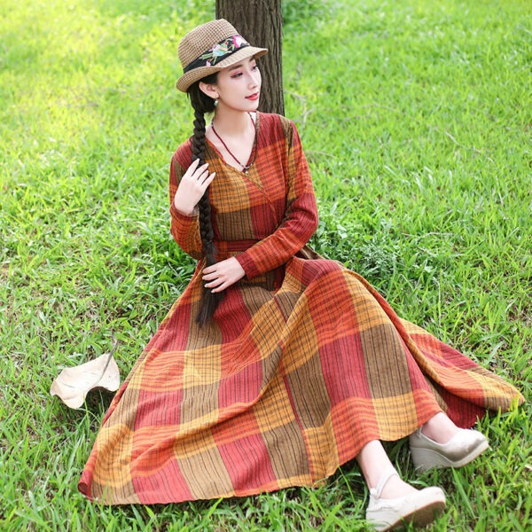 Woman sitting on grass wearing a long plaid dress in warm colors, paired with a straw hat and stylish shoes, enjoying a sunny outdoor setting.