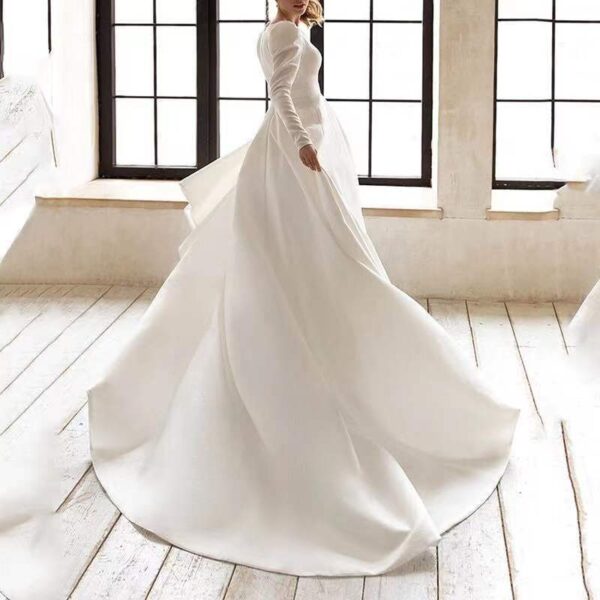 Elegant bride in a flowing white wedding dress with long sleeves, captured in a sunlit room with large windows, showcasing the dress's delicate fabric and graceful silhouette.