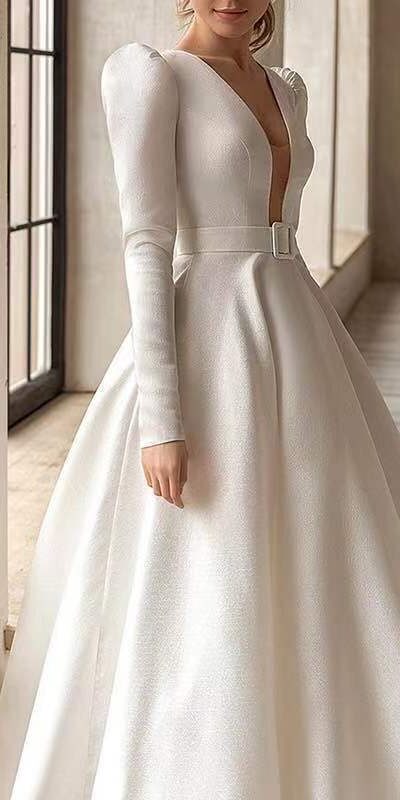 Elegant bride in a stunning white wedding dress with a deep V neckline, long puff sleeves, and a cinched waist, standing gracefully in a bright, modern interior.