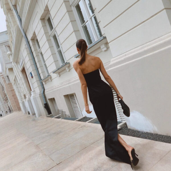 A woman in a strapless black dress walks along a city sidewalk, carrying a black clutch, with a stylish urban building in the background.