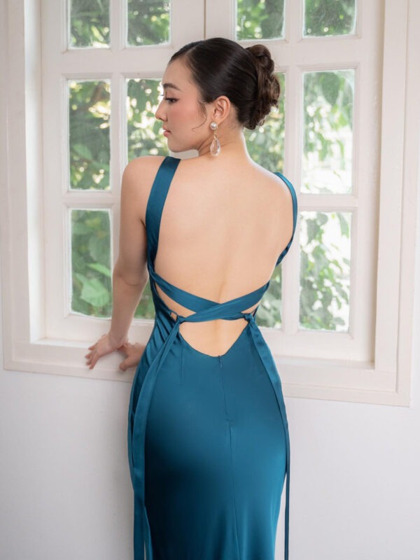 Elegant woman in a backless teal evening gown with intricate cross-strap detailing, posing by a window surrounded by greenery. The dress features a sleek silhouette and is complemented by statement earrings, showcasing a sophisticated style.