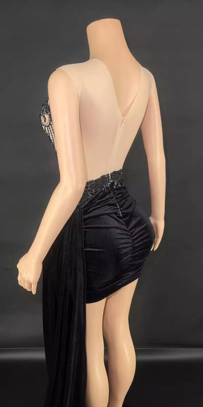 Elegant black evening dress with a sheer back and embellished details, showcased on a mannequin against a dark background. The dress features a fitted silhouette with a draped skirt and intricate beadwork on the bodice.