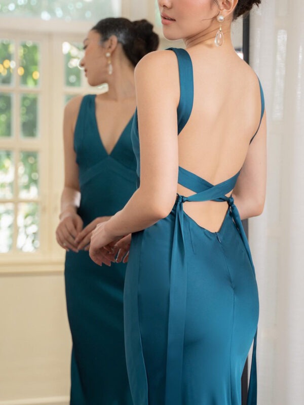 Elegant woman in a teal satin dress with a deep V-back and crisscross straps, admiring her reflection in a mirror. The setting features soft natural light and a stylish interior.