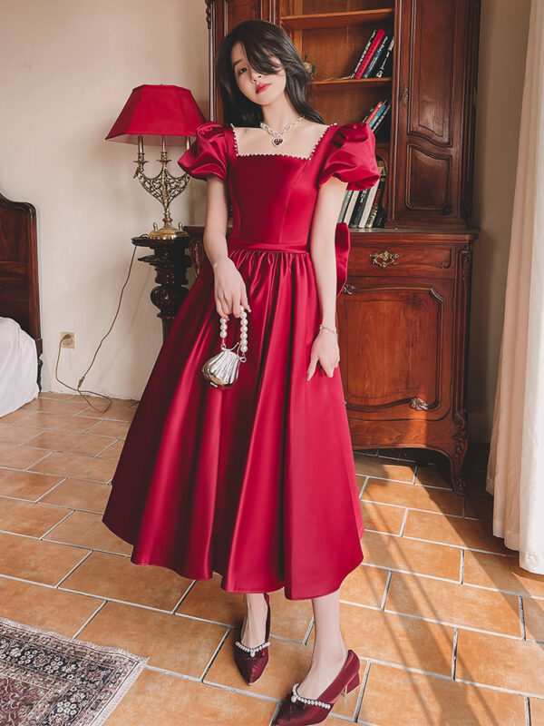 A woman wearing a stylish red puff-sleeve dress stands elegantly in a well-lit room with vintage decor. The dress features a square neckline and a flowing skirt, complemented by matching red shoes adorned with pearls. She holds a small purse and poses confidently against a backdrop of wooden furniture and warm lighting.