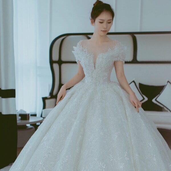 A woman in an elegant, shimmering white ball gown with intricate lace detailing and a deep V-neckline, standing gracefully in a bright, modern room with stylish decor.