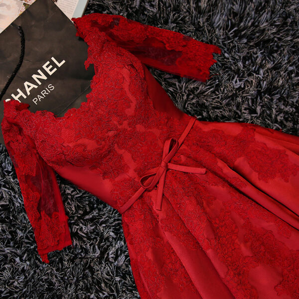 Elegant red lace dress with long sleeves and a bow detail, placed on a textured surface next to a Chanel Paris shopping bag. Perfect for formal occasions and special events.