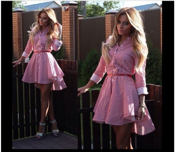 Young woman standing by a wooden fence, wearing a stylish red and white striped dress with a belted waist and flared hem. She has long, wavy blonde hair and is accessorized with a watch and colorful high-heeled shoes. The background features greenery and a sunny outdoor setting.