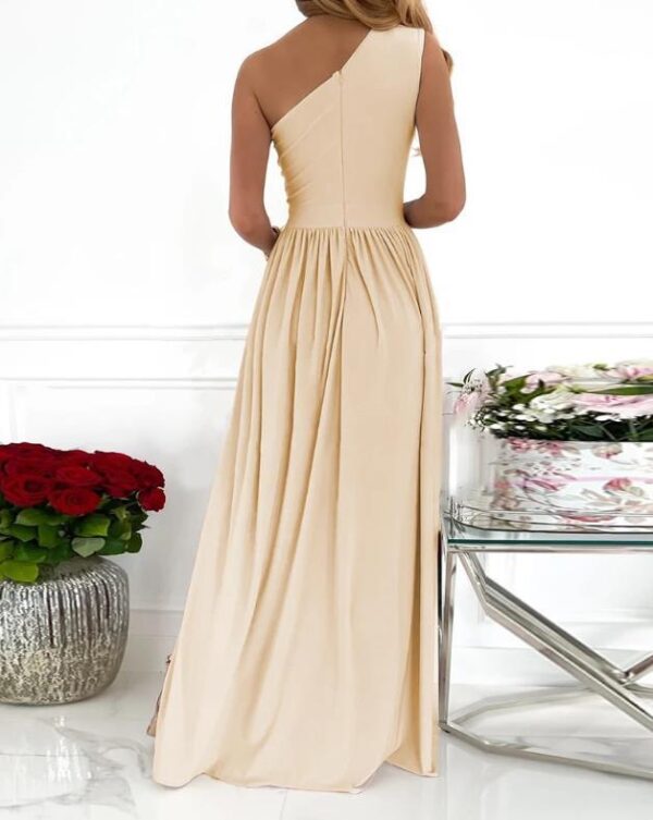 Elegant one-shoulder beige evening gown featuring a flowing design and pleated skirt, showcased against a backdrop of red roses and floral arrangements.