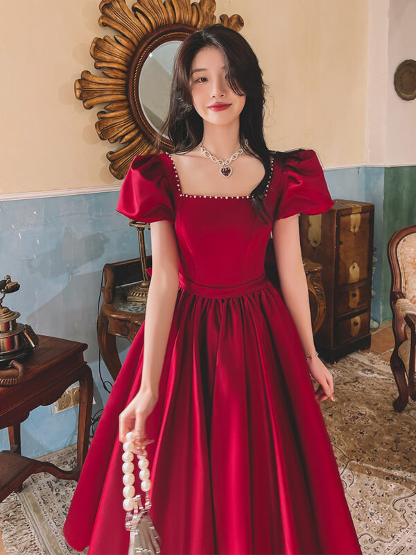 A young woman wearing a stylish red dress with puff sleeves stands in a vintage-inspired room, showcasing elegant jewelry and a classic interior design. The background features a decorative mirror and antique furniture, enhancing the overall aesthetic.