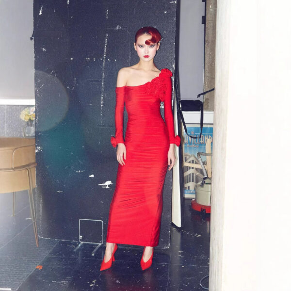 Model in a form-fitting red dress with one shoulder and floral embellishments, posing against a dark backdrop in a stylish indoor setting. The outfit is complemented by matching red heels, showcasing a modern fashion aesthetic.