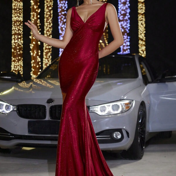 A woman in a stunning red evening gown with a fitted silhouette poses confidently in front of a sleek silver car, illuminated by festive lights in the background. The dress features a deep neckline and sparkling accents, perfect for formal occasions.