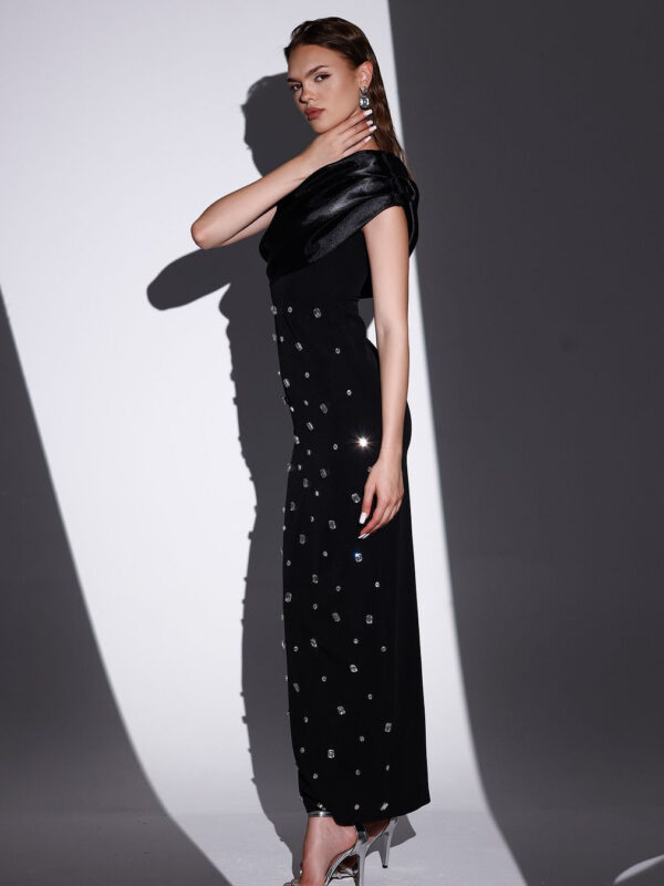 Elegant model posing in a black evening gown adorned with sparkling embellishments, showcasing a sophisticated off-shoulder design and stylish silver heels, set against a dramatic shadow background.