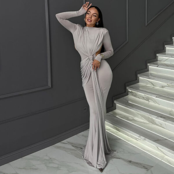 Elegant woman posing in a form-fitting, light gray evening gown with long sleeves and intricate draping details, standing beside a stylish staircase with marble steps and ambient lighting.