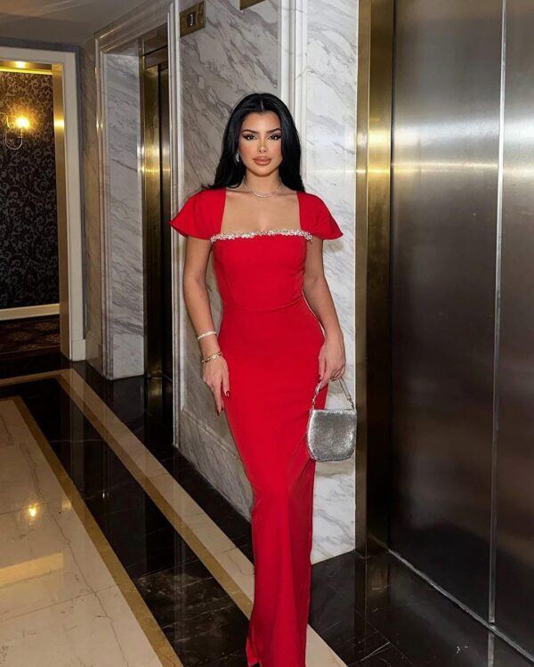 Elegant woman in a red evening gown with embellished neckline, posing confidently in a luxurious hotel hallway near an elevator, showcasing her stylish look and silver handbag.