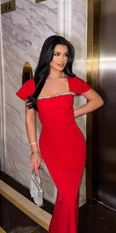 A woman in a stylish red evening dress with short sleeves and embellishments stands in front of an elevator, holding a sparkling clutch purse. The luxurious setting features marble walls and polished flooring.