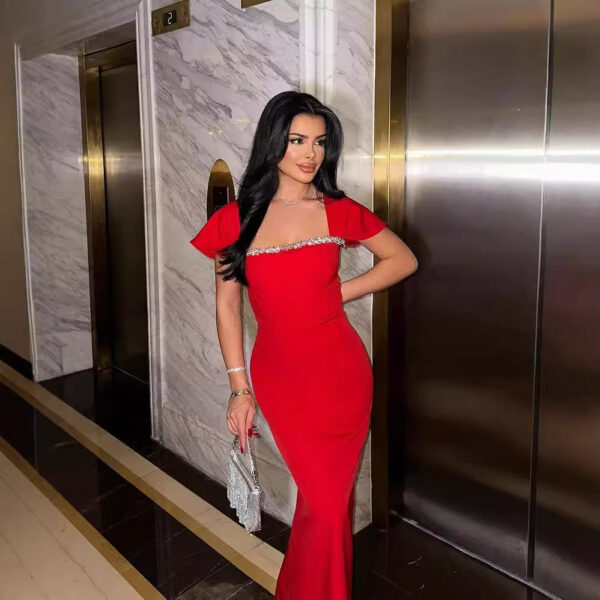 A woman in a stylish red evening dress with short sleeves and embellishments stands in front of an elevator, holding a sparkling clutch purse. The luxurious setting features marble walls and polished flooring.