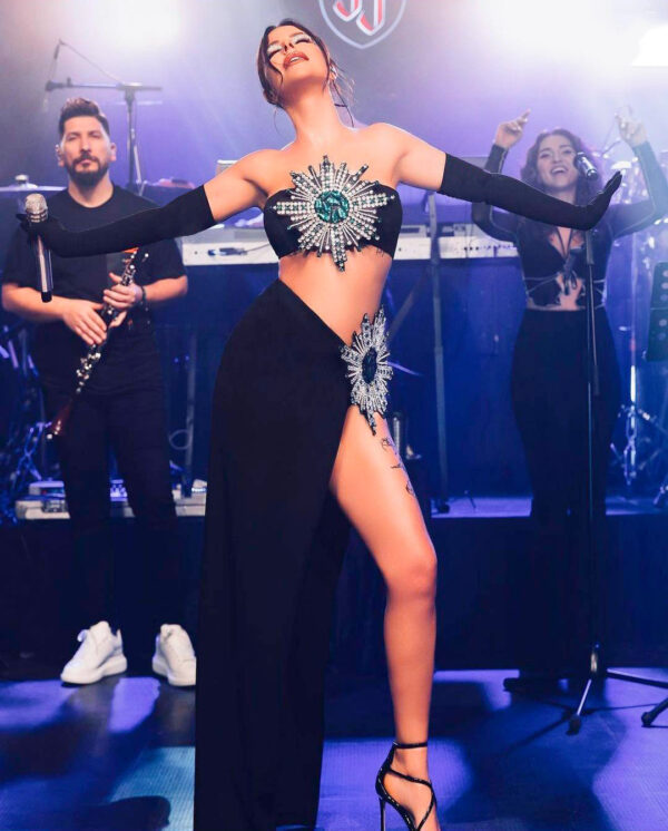 A female singer performs on stage wearing a glamorous black outfit adorned with sparkling embellishments, showcasing a confident pose. Two musicians accompany her, one playing a clarinet and the other singing, with vibrant stage lighting enhancing the energetic atmosphere of the live performance.