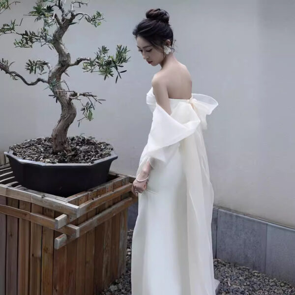 A woman in an elegant off-shoulder white gown stands gracefully beside a bonsai tree, showcasing a serene outdoor setting. The dress features flowing sleeves and a delicate bow at the back, enhancing its sophistication.