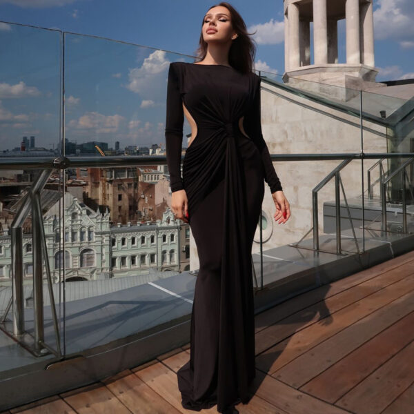 Female model in a stylish black evening gown with cut-out details, posing on a rooftop terrace with a city skyline in the background. The dress features a draped design and long sleeves, highlighting a modern and elegant look against a bright blue sky.