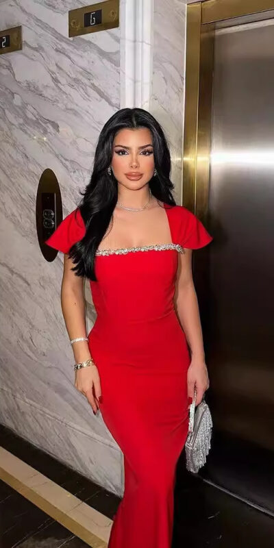 Young woman in a stunning red evening gown with embellished detailing, standing confidently near an elevator in an upscale building, showcasing her elegant style and fashion sense.