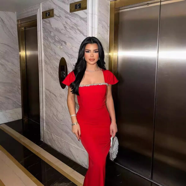 Young woman in a stunning red evening gown with embellished detailing, standing confidently near an elevator in an upscale building, showcasing her elegant style and fashion sense.