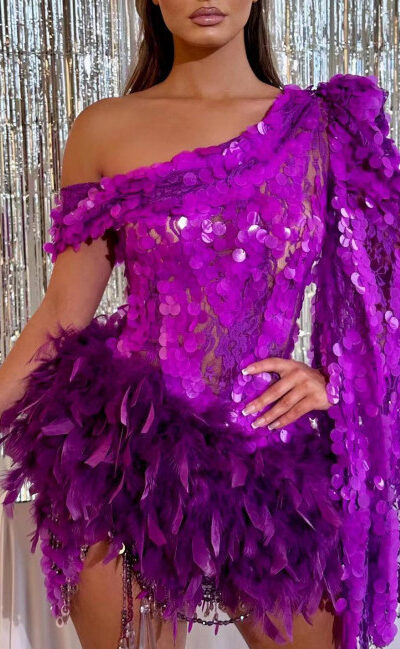 A woman wearing a vibrant purple dress adorned with sequins and feathers, posing elegantly in front of a shimmering silver backdrop, showcasing a stylish one-shoulder design.