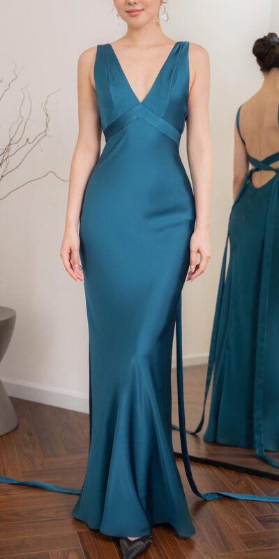 Elegant teal evening gown featuring a deep V-neckline and a fitted silhouette. The dress has a stylish open back with crisscross detailing and flowing ribbons. Styled with minimal accessories, the model showcases the gown in a well-lit room with modern decor.