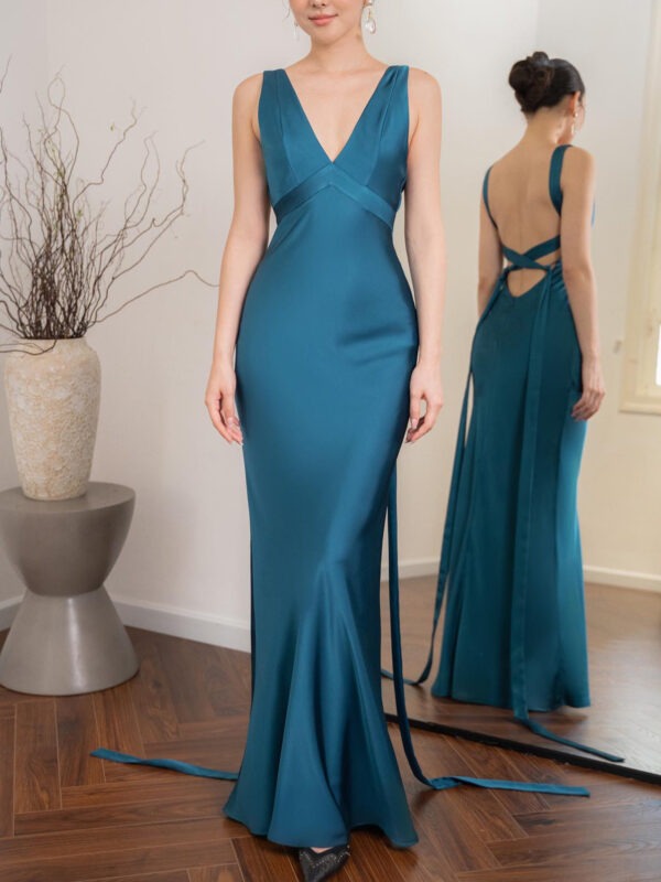 Elegant teal evening gown featuring a deep V-neckline and a fitted silhouette. The dress has a stylish open back with crisscross detailing and flowing ribbons. Styled with minimal accessories, the model showcases the gown in a well-lit room with modern decor.