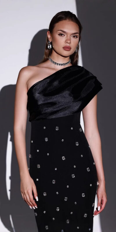 Model wearing a stylish one-shoulder black dress adorned with sparkling embellishments, complemented by elegant jewelry, against a minimalist backdrop.