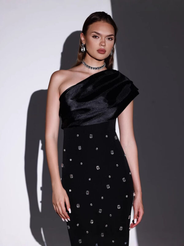 Model wearing a stylish one-shoulder black dress adorned with sparkling embellishments, complemented by elegant jewelry, against a minimalist backdrop.