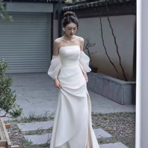 Elegant woman in a white off-the-shoulder wedding dress with puff sleeves, standing gracefully in a serene outdoor setting with stone pathways and greenery. The dress features a sleek silhouette and a thigh-high slit, highlighting modern bridal fashion.