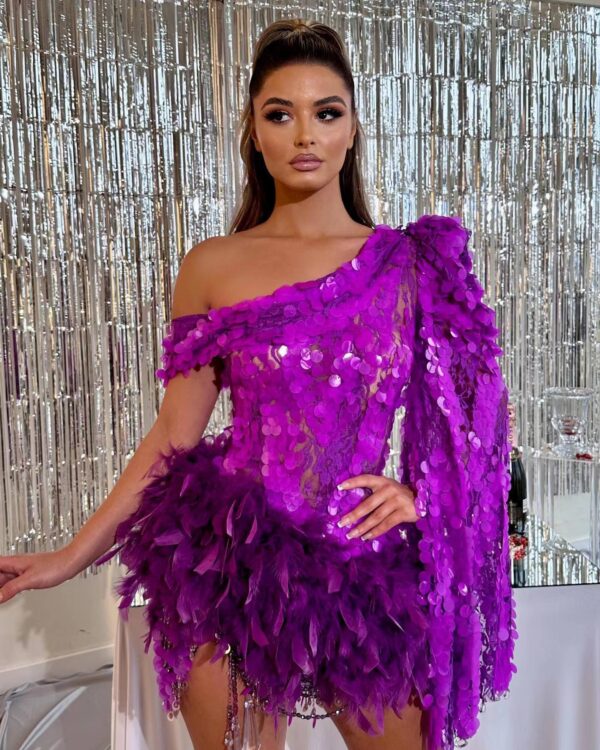 A model poses in a stunning purple dress adorned with sequins and feathers, showcasing a glamorous look against a shimmering silver backdrop. The one-shoulder design highlights her elegance and style, making it perfect for a high-fashion event.