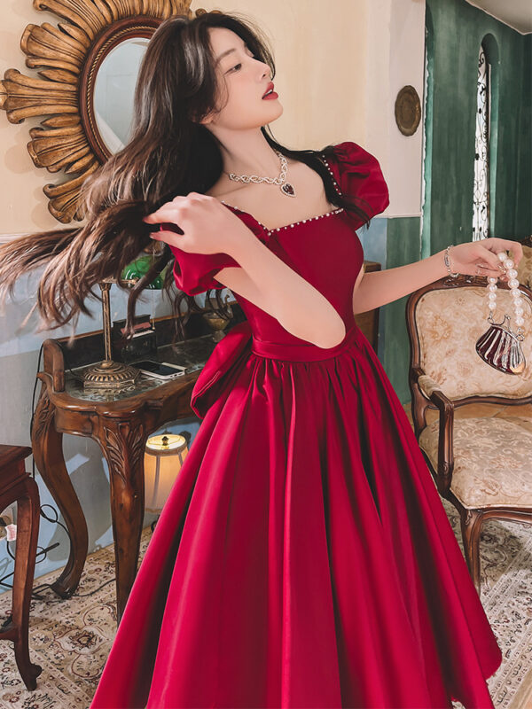 Young woman in a stylish red dress with puff sleeves, posing elegantly while playing with her hair. She accessorizes with a pearl necklace and holds a small handbag, set against a vintage interior with ornate furnishings.