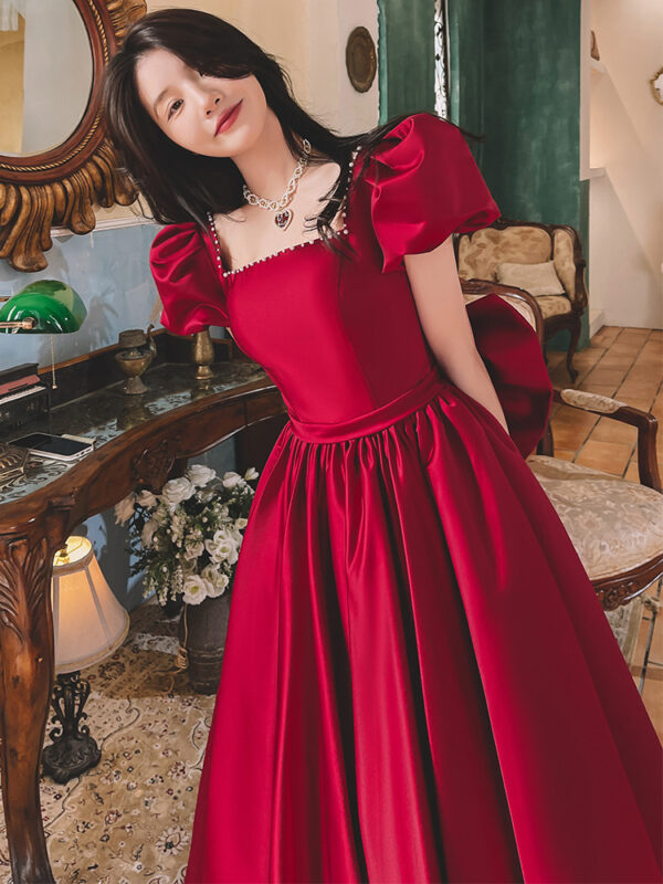 A woman poses elegantly in a vibrant red dress with puff sleeves, showcasing a stylish neckline and a fitted bodice. The setting features vintage decor, including a wooden table, a lamp, and floral arrangements, creating a charming atmosphere.