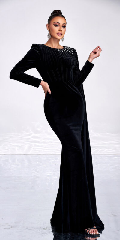 Elegant woman in a black velvet gown with long sleeves and embellished neckline, posing confidently against a minimalist background with reflective surfaces and rocks. Perfect for fashion and evening wear inspiration.