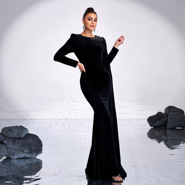 Elegant woman in a black velvet gown with long sleeves and embellished neckline, posing confidently against a minimalist background with reflective surfaces and rocks. Perfect for fashion and evening wear inspiration.