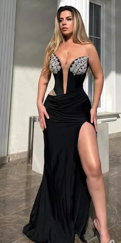 Elegant woman posing in a stunning black evening gown with floral embellishments, featuring a plunging neckline and high slit, showcasing her style and confidence in a modern indoor setting.