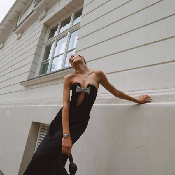 Elegant woman in a strapless black dress with a silver bow detail, posing against a textured wall, showcasing a chic fashion style.