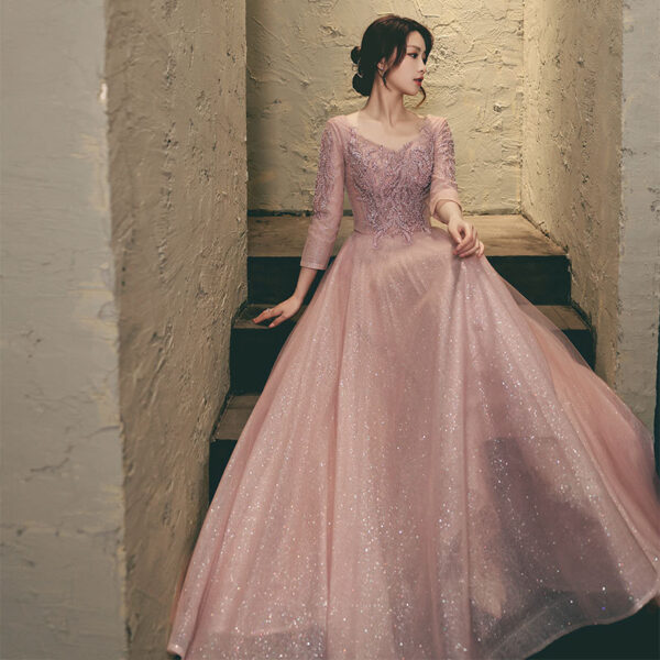 Elegant woman in a sparkling pink gown with intricate lace details, sitting on a staircase against a textured wall. The dress features three-quarter sleeves and a flowing skirt, perfect for formal occasions or evening events.