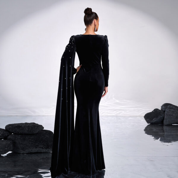 Elegant woman in a black velvet gown with a dramatic long sleeve and embellished details, standing against a serene backdrop of smooth water and dark rocks, showcasing a sophisticated fashion statement.