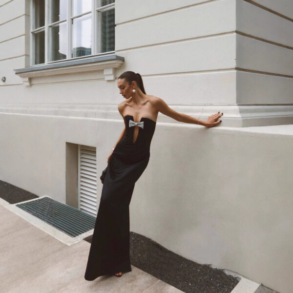 A model poses elegantly against a minimalist wall, wearing a stylish black strapless gown with a silver bow detail at the waist. The setting features large windows and a modern architectural design, emphasizing contemporary fashion aesthetics.