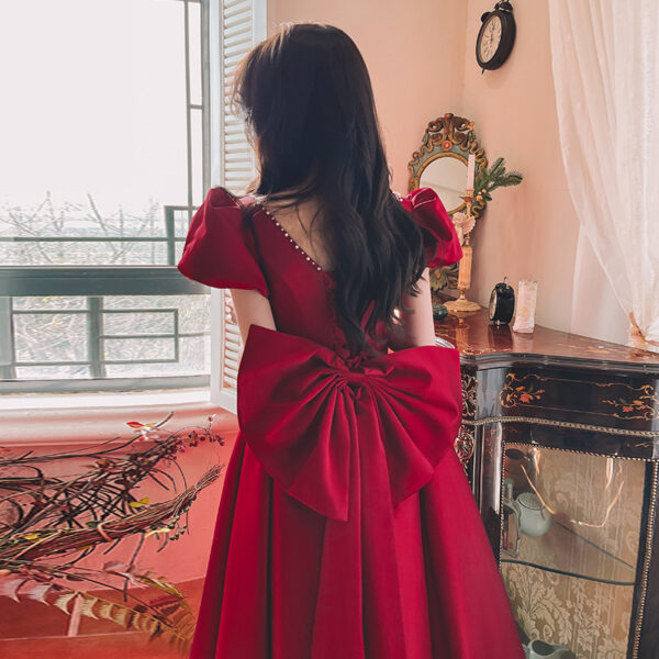 A woman wearing a red dress with puff sleeves and a large bow at the back, standing in a well-lit room with vintage decor and a window in the background.