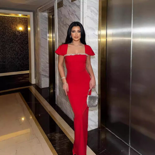 Elegant woman in a red evening gown with jeweled neckline, standing confidently in a luxurious hotel hallway next to an elevator.