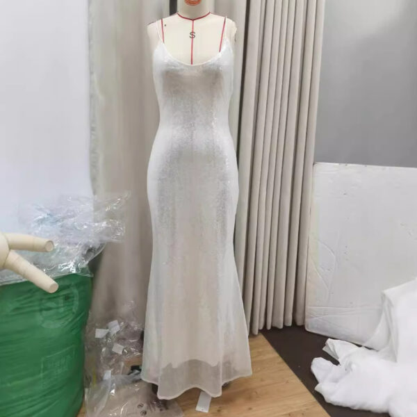 Elegant white evening gown displayed on a mannequin, featuring a fitted silhouette and delicate spaghetti straps, set against a neutral backdrop with fabric drapes and packaging materials.