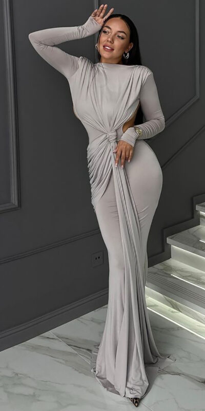 Elegant woman in a form-fitting, long-sleeve beige dress with a twisted front design, posing confidently against a stylish gray wall and modern staircase with illuminated steps.