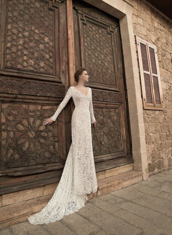 Elegant bride in a long-sleeved lace wedding dress poses against ornate wooden doors, showcasing intricate detailing and a flowing train, set in a charming stone backdrop. Perfect for bridal fashion inspiration.